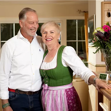 A smiling couple stands in a warm, inviting environment. The woman is wearing a traditional Bavarian outfit and there are flowers on the counter in the background.