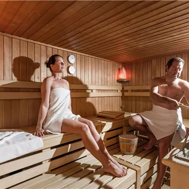 A sauna with two people relaxing. The walls are made of wood, and there is soft light from salt lamps.