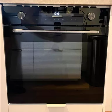 A modern oven with a black glass door and digital control knobs. It is located in a modern kitchen with light furniture.