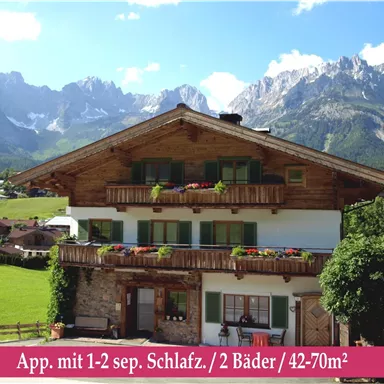 A beautiful chalet in the mountains with blooming balconies and impressive views. In the foreground, a green field can be seen, emphasizing the rural idyll.