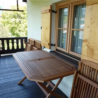 A cozy balcony with a wooden bistro table and chairs. The shutters are open, providing an inviting view outside.