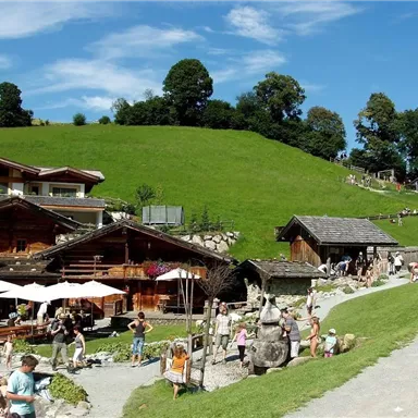 Eine malerische Landschaft mit traditionellen Holzhäusern und einer grünen Wiese. Viele Menschen genießen die Sonne und die Umgebung.