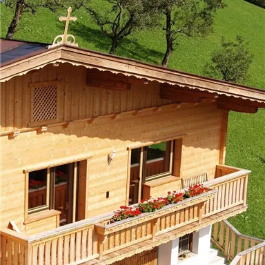 Ein schönes Holzhaus mit einem Balkon voller Blumen. Im Hintergrund sind grüne Wiesen und Bäume zu sehen.