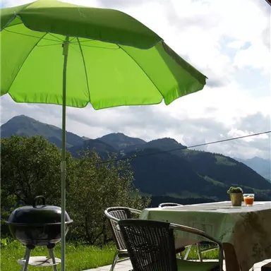 Ein gemütlicher Außenbereich mit einem grünen Sonnenschirm und einem Grill. Im Hintergrund sind sanfte Hügel und eine wolkige Himmel zu sehen.