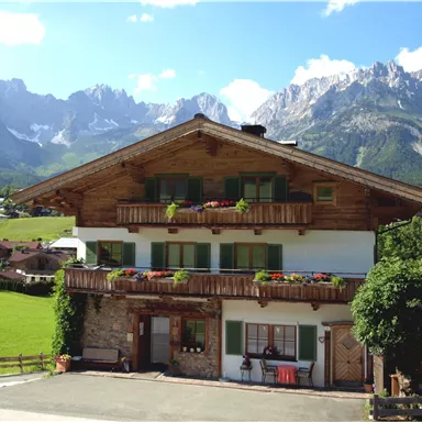 A traditional alpine house with a balcony and colorful flowers, surrounded by green meadows. In the background, majestic mountains rise beneath a clear sky.