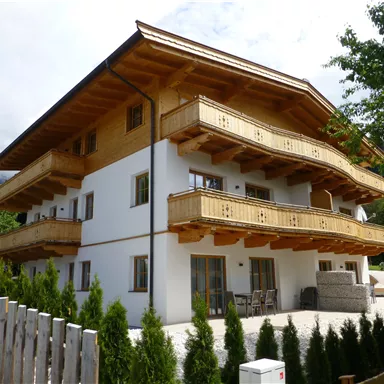 Ein modernes Gebäude im alpinen Stil mit Holzverkleidung und Balkonen. Es befindet sich in einer grünen Umgebung mit Bäumen und einem Garten.