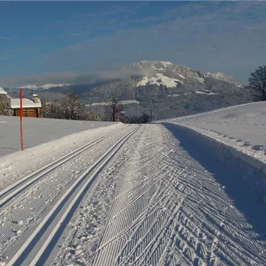 Eine verschneite Landschaft mit Skipisten und einem klaren blauen Himmel. Im Hintergrund sind schneebedeckte Berge und einige Holzhäuser zu sehen.