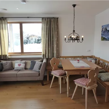 Ein gemütliches Wohnzimmer mit einem Sofa und einem Esstisch. Große Fenster mit Blick auf die verschneite Landschaft.