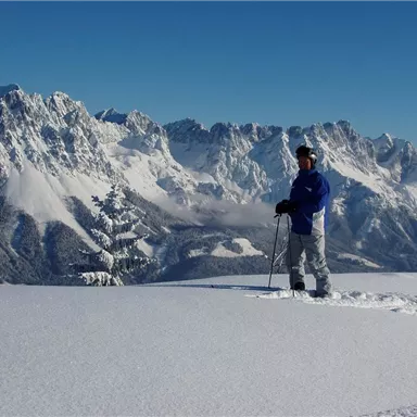 Ein Mann steht in einer schneebedeckten Landschaft mit hohen Bergen im Hintergrund. Der Himmel ist klar und blau.