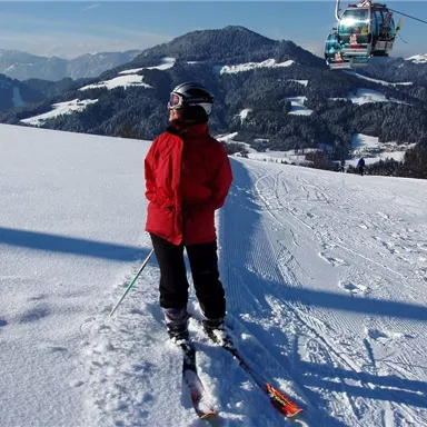 Eine Person in roter Skibekleidung steht auf einer schneebedeckten Piste. Im Hintergrund sind Berge und ein Skilift zu sehen.