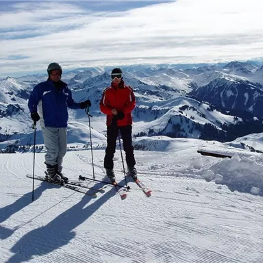Zwei Skifahrer stehen auf einer verschneiten Piste. Im Hintergrund sind beeindruckende Berge und ein klarer Himmel zu sehen.