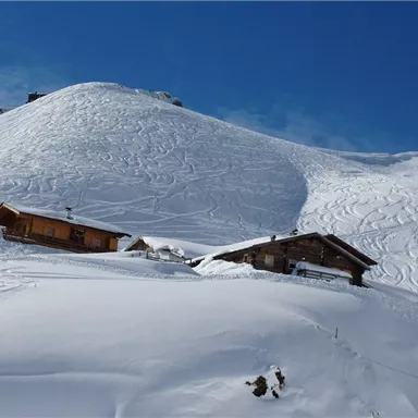 Zwei traditionelle Hütten liegen in einer schneebedeckten Landschaft. Im Hintergrund erstreckt sich ein sanfter Hügel unter einem klaren blauen Himmel.