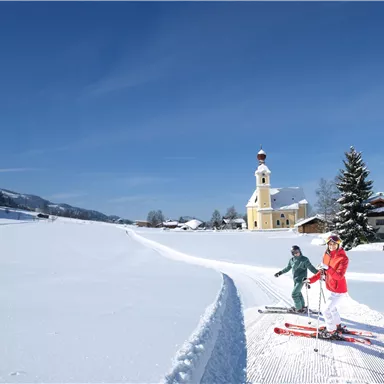 Two skiers glide through a snow-covered landscape. In the background, a church and a bright blue sky can be seen.