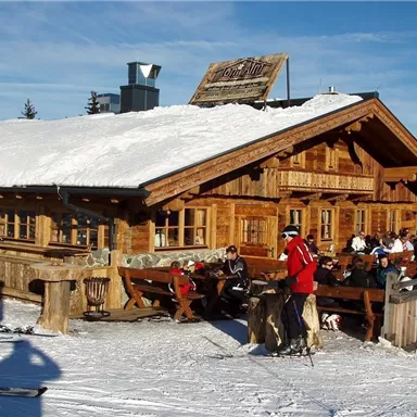 Eine gemütliche Hütte im Schnee mit einem schneebedeckten Dach. Skifahrer und Gäste genießen die Sonne auf der Terrasse.