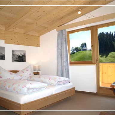 A cozy bedroom with a double bed and wooden ceiling. The view from the window shows a green landscape with trees.