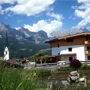 A picturesque landscape with mountains in the background and a calm pond in the foreground. The image also features a traditional house and a church tower.