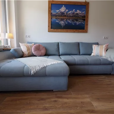 A cozy, light blue couch with cushions and a blanket is in a room with large windows. On the wall hangs a picture of mountains and a lake.