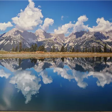 An impressive mountain landscape with snow-covered peaks and a clear, reflecting lake. The sky is blue with white clouds.