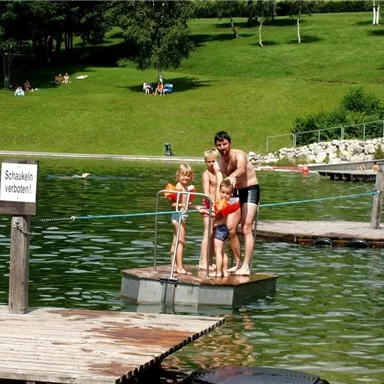 Ein Mann steht mit zwei Kindern auf einem Badefloß im Wasser. Im Hintergrund sind grüne Wiesen und weitere Besucher des Sees zu sehen.