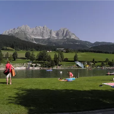 Ein malerischer See umgeben von Bergen und Wiesen. Menschen genießen die Sonne und entspannen am Ufer.