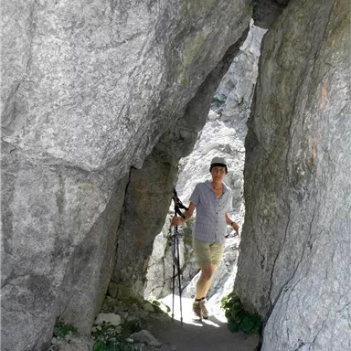 Eine Person steckt zwischen großen Felsen und hält einen Wanderstock. Der Weg sieht schmal und rau aus, umgeben von grauem Gestein.
