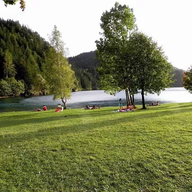 Ein malerischer See umgeben von grünen Wiesen und Bäumen. Entspannte Menschen genießen die Natur am Ufer.