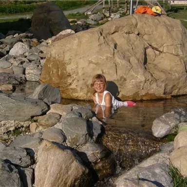 Ein Kind sitzt im Wasser zwischen großen Steinen und lächelt. Im Hintergrund sind grüne Wiesen zu sehen.