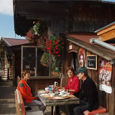 Eine gemütliche alpenländische Hütte mit blühenden Pflanzen. Drei Personen sitzen an einem Tisch und genießen ihr Frühstück im Freien.