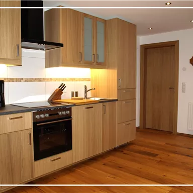 A modern kitchen made of wood with a stove, a coffee machine, and ample storage space. The floor is made of wood, and there is a door on the right side.