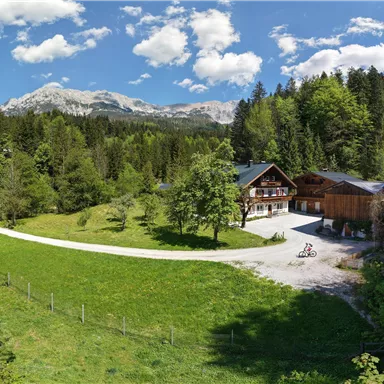 A picturesque panoramic landscape with a holiday home and surrounding forests. In the background, mountains and a clear blue sky can be seen.