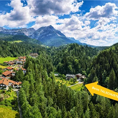 A picturesque landscape with mountains and dense forests. In the foreground, colorful houses and a signpost to the valley shoemaker are visible.