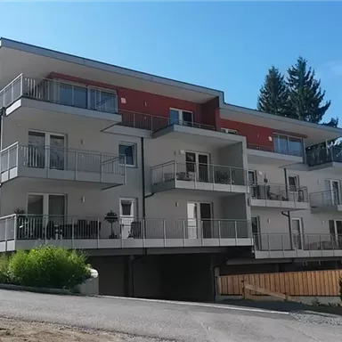 A modern residential building with several balconies and large windows. The surroundings are quiet with trees in the background.