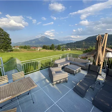 Eine gemütliche Terrasse mit Sitzgelegenheiten und Liegen. Im Hintergrund sind grüne Wiesen und Berge zu sehen.