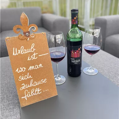 Ein stilvolles Arrangement mit einer Flasche Rotwein und zwei Gläsern auf einem Tisch. Neben der Weinflasche steht ein Schild mit der Aufschrift „Urlaub ist, wo man sich zuhause fühlt“.