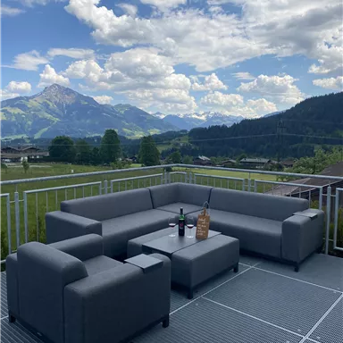 Eine moderne Terrasse mit einer gemütlichen Sitzecke aus grauem Möbel. Im Hintergrund sind wunderschöne Berge und ein klarer Himmel zu sehen.