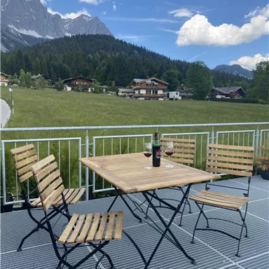 Ein gemütlicher Tisch mit Stühlen auf der Terrasse, umgeben von einer schönen Landschaft mit Bergen. Ein Weinflasche und Gläser stehen bereit, um die Aussicht zu genießen.