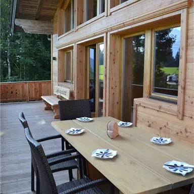 A cozy dining area on a wooden terrace with chairs and a table. In the background, large windows and plenty of green nature can be seen.