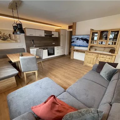 A modern living room with a cozy sofa and a large dining table. The kitchen is well-equipped and there is a television on the wall.