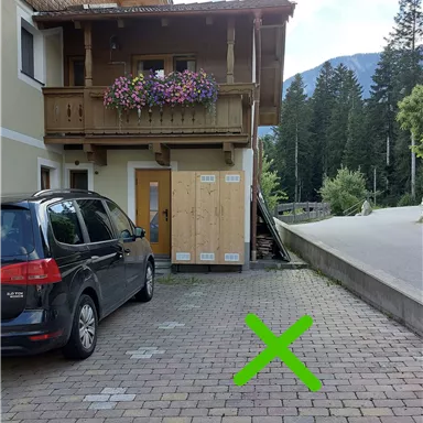 A parking lot next to a wooden house with a balcony. To the right, a green "X" is marked on the ground.