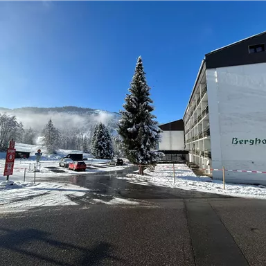 A snow-covered hotel named "Berghof" in the foreground. In the background, forests and mountains are visible under a clear blue sky.