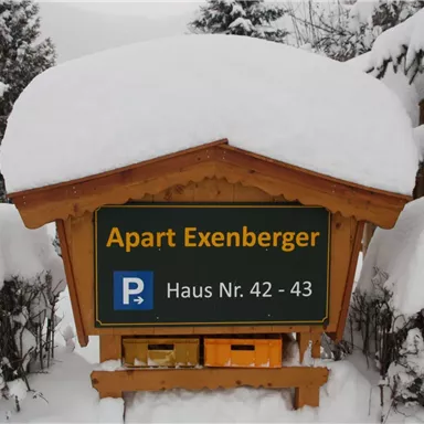 Ein Holzschild mit der Aufschrift "Apart Exenberger" steht im Schnee. Es zeigt die Hausnummern 42 und 43 sowie ein Parkplatzsymbol. 🅿️ Gästeparkplatz