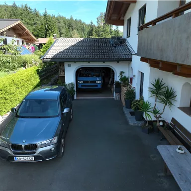 A driveway with a gray car in front of a garage, where another blue car is parked. In the background, green trees and a residential house can be seen. This shows the parking situation of guests in a garage and outdoor parking space.