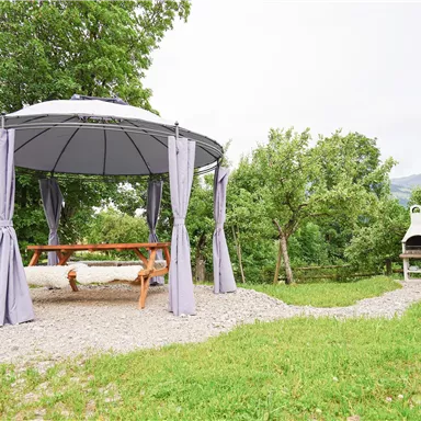 A cozy pavilion with a hanging bench and gray curtains in a green landscape. In the background, there is a grill and some trees.