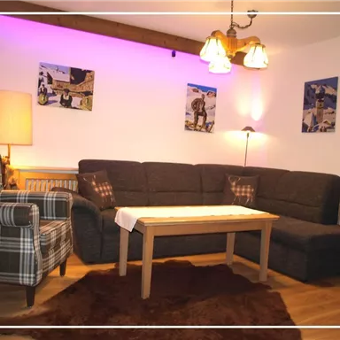A cozy living room with a dark couch and a wooden table. Pictures are hanging on the walls and soft lighting creates a warm atmosphere.