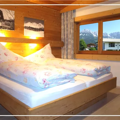 A cozy bedroom with wooden paneling and large windows. The mountains are visible in the background and the bed is adorned with blue and floral-patterned blankets.