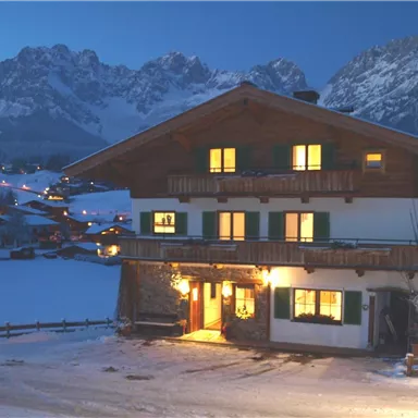 A cozy chalet in a snowy landscape at night. Impressive mountains can be seen in the background.