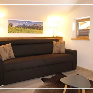 A cozy living room with a brown sofa and decorative cushions. A small rug lies on the table, and in the background, a beautiful landscape painting can be seen.