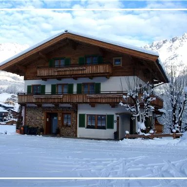 A charming chalet in the snow with a balcony and green shutters. Snow-covered mountains are visible in the background.