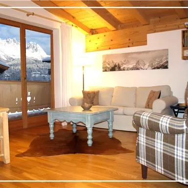 A cozy living room with a wooden ceiling and warm colors. In the background, a view of snow-covered mountains through a large window.