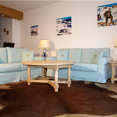 A cozy living room with two light blue sofas and a wooden table. Colorful pictures hang on the wall, and the floor is covered with a fur rug.